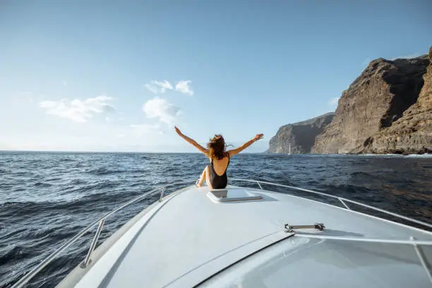 Woman enjoying ocean voyage sitting back on the yacht nose while sailing near the breathtaking rocky coast on a sunset. Wide seascape view