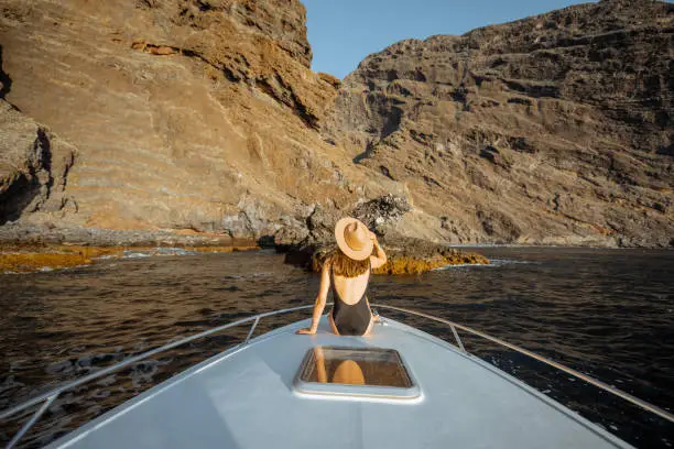 Woman in swimsuit and sun hat enjoying ocean voyage sitting on the yacht nose while sailing near the breathtaking rocky coast on a sunset. Wide seascape view