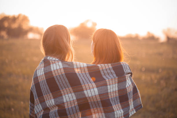 Two young women covered by blanket looking on the sunset. Best friends
