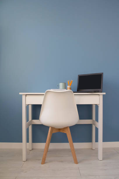 istock Front view of empty home office of creative entrepreneur with white table during lunch break. Modern creative workspace with black laptop, cup of tea and yellow pencils in paper tube. 1207585624