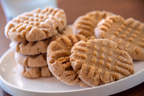 Horizontal image of a plate of homemade peanut butter cookies sitting on a kitchen table with window light streaming in from behind. Focus is on cookies in the foreground and using shallow depth of field the focus fades quickly.