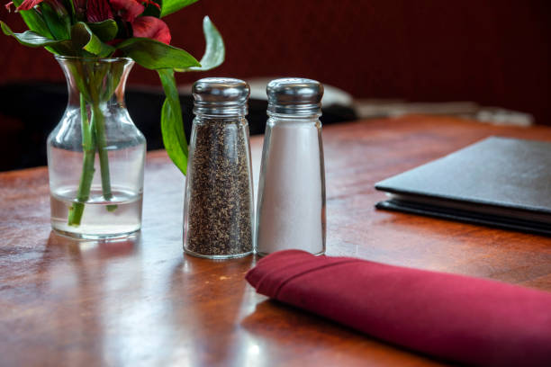 Horizontal image of a salt and pepper shaker sitting on a restaurant table. There is a red cloth napkin, menu and flowers in a vase sitting on the wooden table.
