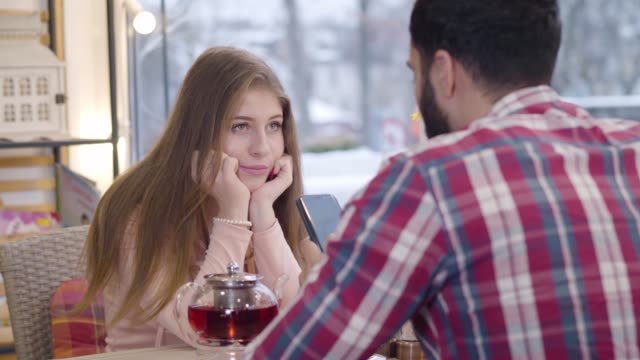 Portrait of bored pretty Caucasian girl sitting with boyfriend or husband in restaurant as her spouse using smartphone. Bearded Middle Eastern man ignoring beautiful partner. Dating, disappointment.