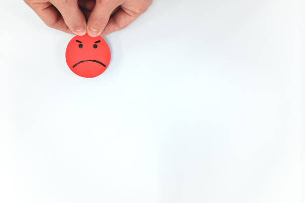 istock Hand holding a red angry emoticon in white background with copy space. Anger, bad customer service and unsatisfied feedback evaluation. 1207616412