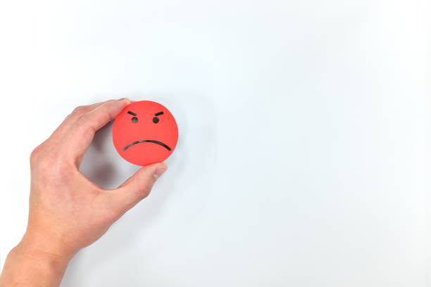 istock Hand holding a red angry emoticon in white background with copy space. Anger, bad customer service and unsatisfied feedback evaluation. 1207616517