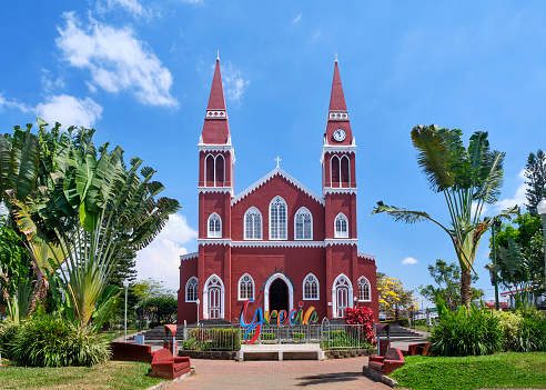 Grecia, Costa Rica - February 11, 2020: The all metal red and white catholic church in Grecia Costa Rica, sits between green palm trees and under a beautiful blue sky with white fluffy clouds.