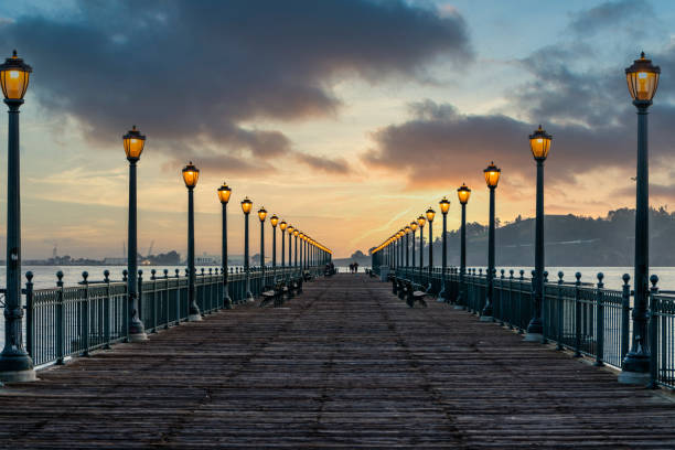 Dramatic sunrise over the San Francisco Bay from Pier 3 lined with illuminated lamp lights on the Embarcadero. Treasure Island is visible in the background.