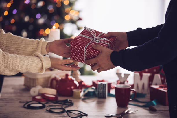 Woman giving a Christmas gift to man Shot of unrecognizable woman giving a Christmas gift box to a man in front of a wooden table with abundance of Christmas themed decorations and ribbons. gift lounge stock pictures, royalty-free photos & images