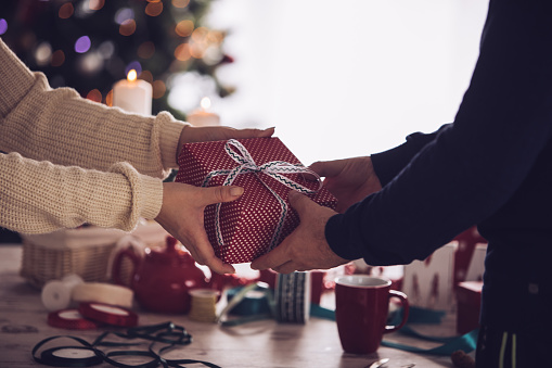 Shot of unrecognizable heterosexual couple sharing a beautifully wrapped Christmas gift in front of a rustic wooden table with ribbons and decoration.