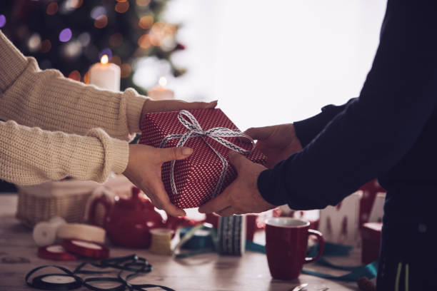 Unrecognizable couple sharing a Christmas gift Shot of unrecognizable heterosexual couple sharing a beautifully wrapped Christmas gift in front of a rustic wooden table with ribbons and decoration. gift lounge stock pictures, royalty-free photos & images