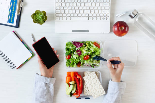 mani di donna d'affari che lavora al telefono e mangia sano pranzo di lavoro sul posto di lavoro. verdure e insalata verde fresca nella scatola del pranzo sulla scrivania con laptop e altre forniture per ufficio. - breakfast salad leaf vegetable foto e immagini stock