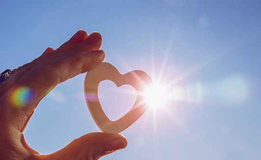 Close up view of woman hand holding white wooden heart against sun and blue sky with lens flare and sun`s photography star effect. Positive emotions concept.