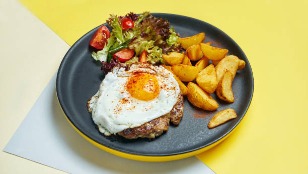 Beef steak with fried eggs with a side dish of salad and fried potatoes on a black plate on a colored background. Appetizing food for lunch stock photo