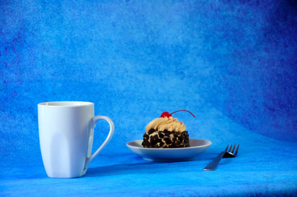 White ceramic mug of hot tea and a plate with chocolate biscuit cake with cherry and cream.