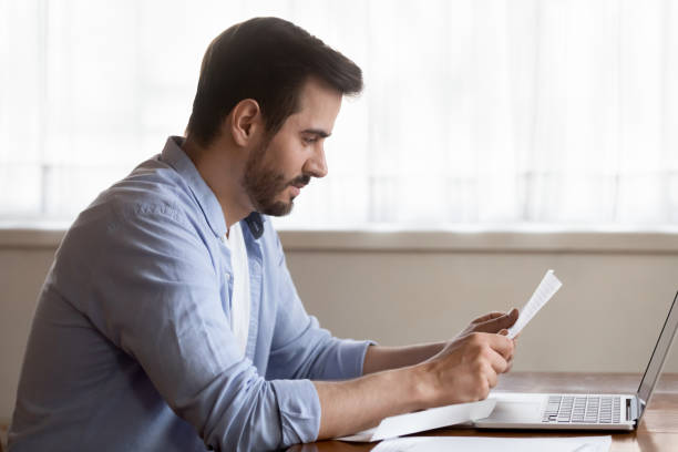 Focused millennial man read letter at home Focused millennial male sit at table at home reading pleasant good news in post letter correspondence, concentrated Caucasian man work on laptop at desk consider paper agreement or contract college acceptance letter stock pictures, royalty-free photos & images