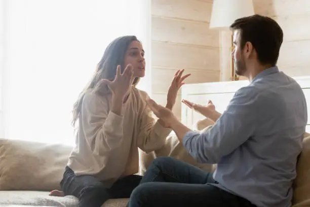 Angry young couple sit on couch in living room having family fight or quarrel suffer from misunderstanding, millennial husband and wife dispute involved in argument, relationships problems concept