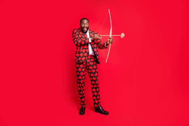 Full body photo of funny dark skin man with bow and love arrow amour cupid role see good couple wear hearts pattern suit shirt necktie tie boots outfit isolated red color background Full body photo of funny dark skin man with bow and love arrow amour, cupid role see good couple wear hearts pattern suit shirt necktie tie boots outfit isolated red color background bow and arrow photos stock pictures, royalty-free photos & images