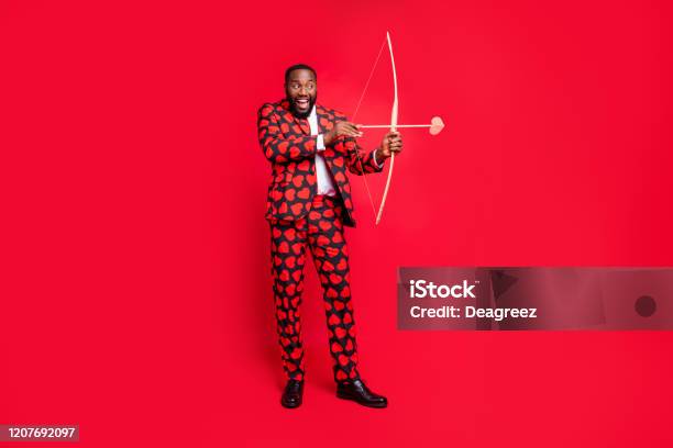 Full Body Photo Of Funny Dark Skin Man With Bow And Love Arrow Amour Cupid Role See Good Couple Wear Hearts Pattern Suit Shirt Necktie Tie Boots Outfit Isolated Red Color Background Stock Photo - Download Image Now