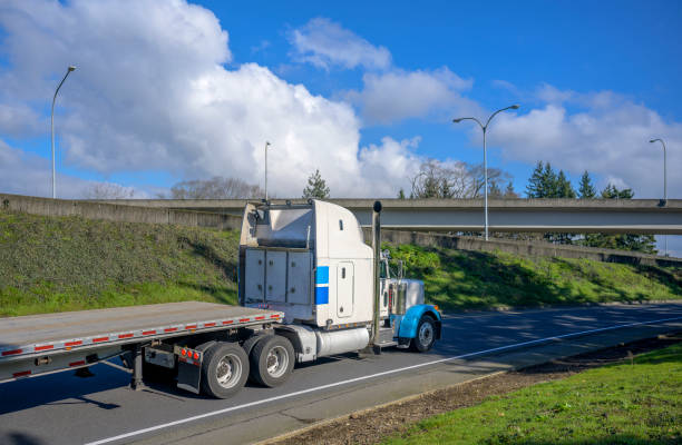 istock Big rig classic high cab semi truck with storage boxes on the back side transporting empty flat bed semi trailer running on the road with bridge ahead 1207698615