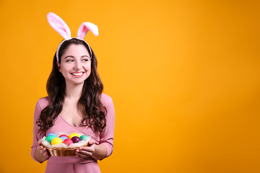 Studio portrait of young beautiful woman wearing traditional bunny ears headband for easter and smiling. Brunette female with wavy hair over yellow background. Close up, copy space.