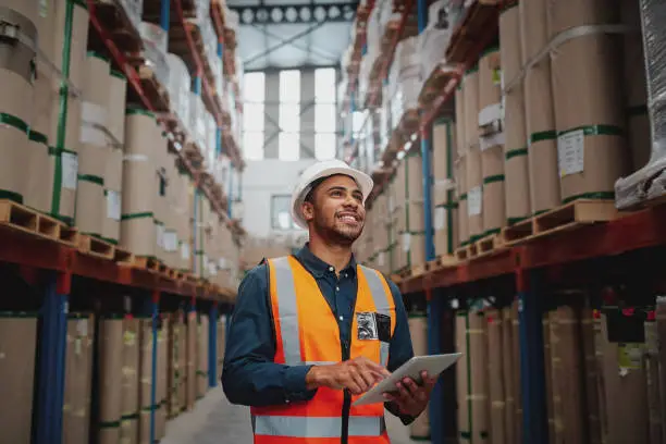 Photo of Low angle view of young african man wearing reflective jacket holding digital tablet standing in factory warehouse smiling