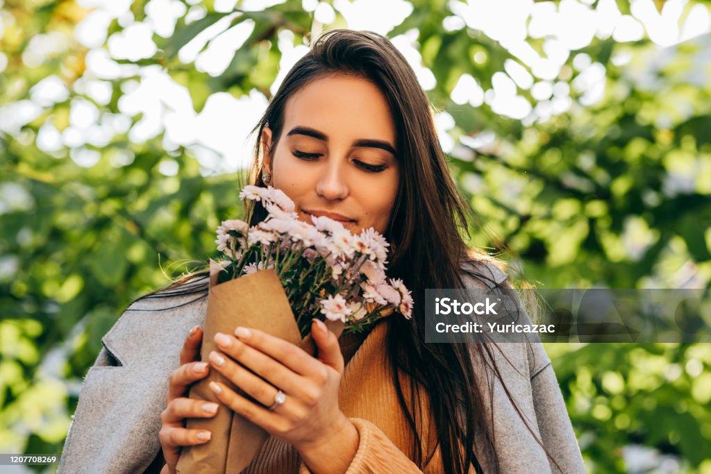 Outdoor close-up portrait of beautiuful young woman smelling the pink flowers on sunny day in the park. Happy female recieved a gift a bouquet of flowers from her boyfreind in the city street. Woman's day 20-29 Years Stock Photo
