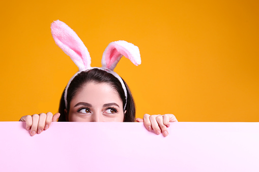 Cropped studio portrait of young beautiful woman wearing traditional bunny ears headband for easter hiding behind pink wall. Brunette female with wavy hair over yellow background. Close up, copy space