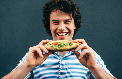 Close-up portrait of young happy man eating a healthy hamburger. Handsome man in a fast food restaurant eating a hamburger outdoors. Man with curly hair having street food and eat a burger.