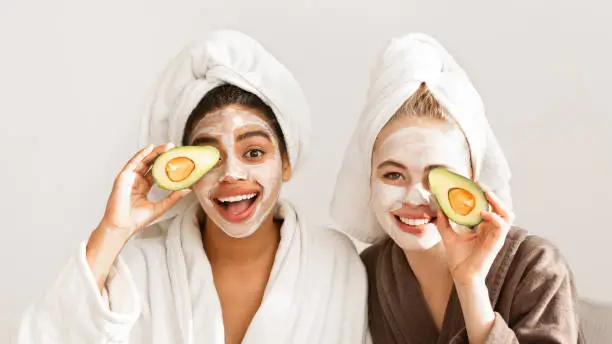 Emotional multiracial girlfriends in bathrobes and face masks on covering eyes with avocado halves, panorama
