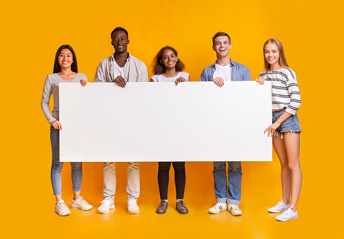 Smiling group of multiracial students standing together in row and displaying white empty placard, yellow studio background