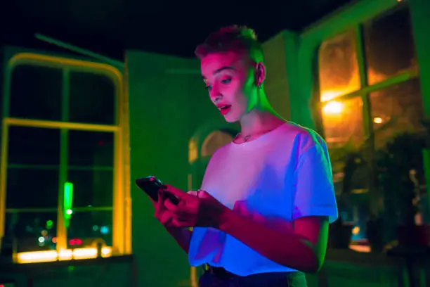 Surprised. Cinematic portrait of stylish woman in neon lighted interior. Toned like cinema effects, bright neoned colors. Caucasian model using smartphone in colorful lights indoors. Youth culture.