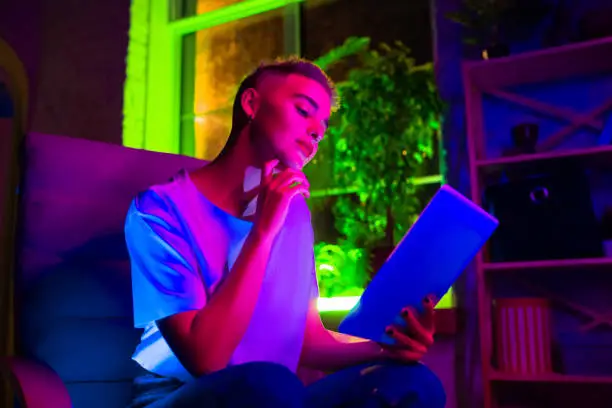 Interested. Cinematic portrait of stylish woman in neon lighted interior. Toned like cinema effects, bright neoned colors. Caucasian model using tablet in colorful lights indoors. Youth culture.