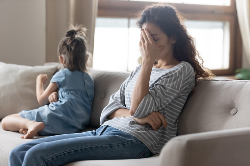 Unhappy young mother touching forehead, feeling tired of bad daughter's behavior at home. Offended little child girl sitting on different side on couch, ignoring sad frustrated mother in living room.