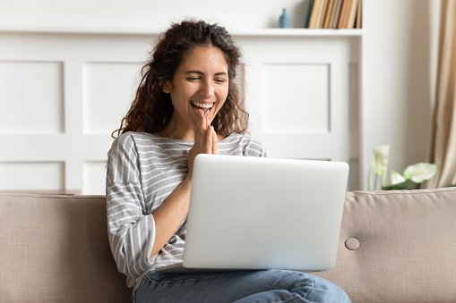 Overjoyed young woman looking at laptop screen, feeling excited about good news at home. Happy attractive millennial lady received online lottery win notification, sitting on sofa in living room.