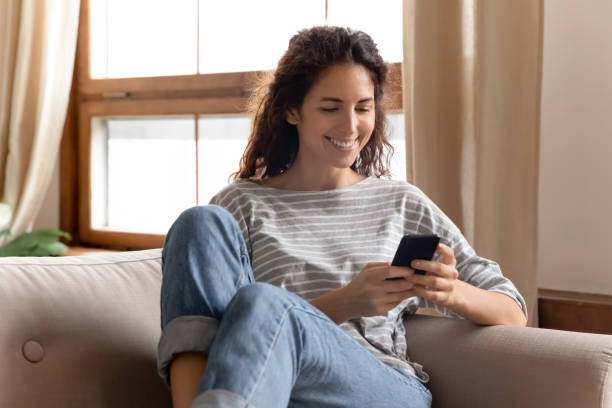 Smiling young lady using mobile applications at home. Happy pleasant millennial woman relaxing on comfortable couch, holding smartphone in hands. Smiling young lady chatting in social networks, watching funny videos, using mobile applications at home. dependency photos stock pictures, royalty-free photos & images