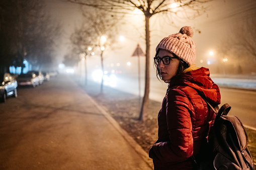 Scared young woman on city street in foggy night looking over shoulder behind.