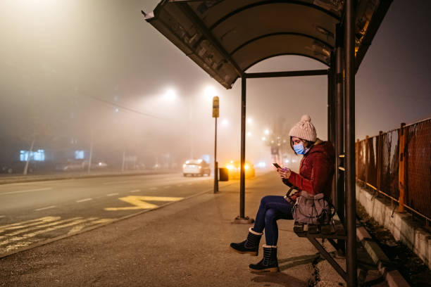 donna con maschera con il telefono alla fermata dell'autobus - global warming smog city pollution foto e immagini stock