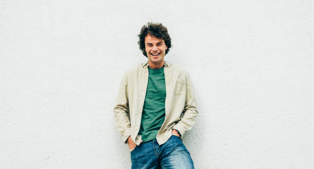 istock Image of handsome young man with curly hair smiling and posing for advertisement, looking at the camera with copy space for your text, standing at building concrete wall outdoors. 1207728152