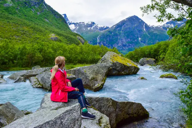 The young woman near river which is located near path to the Briksdalsbreen (Briksdal) glacier. The melting of this glacier forms waterfall and river with clear water. Jostedalsbreen National Park. Norway.