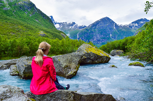 The young woman near river which is located near path to the Briksdalsbreen (Briksdal) glacier. The melting of this glacier forms waterfall and river with clear water. Jostedalsbreen National Park. Norway.