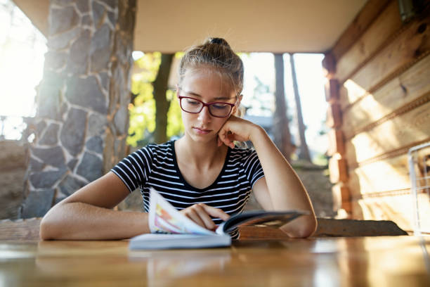 Teenage girl reading a comic book on terrace Teenage girl is sitting on at the wooden table on terrace of a small wooden cabin. The girl is reading a comic book on a sunny morning. reading comic book stock pictures, royalty-free photos & images