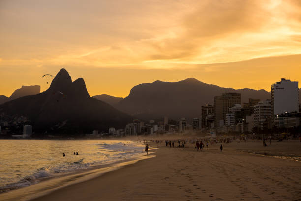 Ipanema beach at sunset in Rio de Janeiro, Brazil Rio de Janeiro, Brazil - July 2nd, 2016: Ipanema beach with silhouette of people relaxing & having fun & a paraglider overhead at sunset & with Dois Irmãos Two Brothers mountains in background and hotels and buildings along the beachfront in Rio de Janeiro, Brazil, South America two brothers mountain stock pictures, royalty-free photos & images