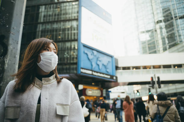 young asian woman wearing a protective face mask to prevent the spread of germs and viruses in the city - hong kong city urban scene building exterior imagens e fotografias de stock