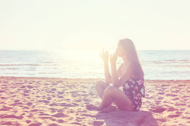 Girl meditates on the beach in summer morning, bright sunlight around.