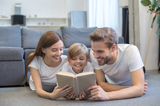 Smiling father and mother lying on floor near their son while reading book at home. Family and happiness concept