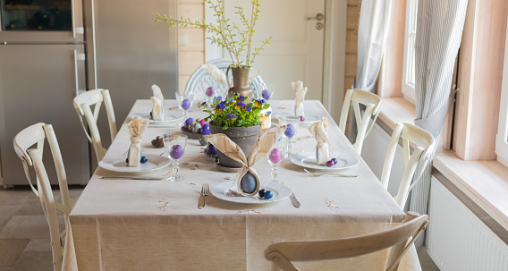 Easter festive spring table setting decoration, bunny ears shaped napkins, dyed hen's and quail eggs, cakes, violets potted, feathers, family dinner or breakfast concept, banner