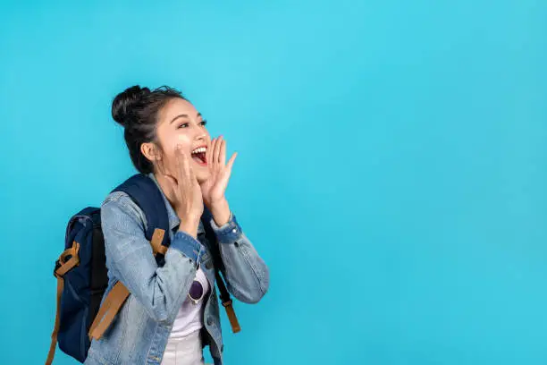 Photo of Happy asian woman travel backpacker shouting open mouth to copyspace on blue background. Cute asia girl smiling wearing casual jeans shirt and looking to aside for present promotions.