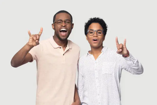Overjoyed mixed race ethnic married couple in glasses having fun together, shouting, singing, showing rock-n-roll gesture, posing for photo, looking at camera isolated on grey studio background.