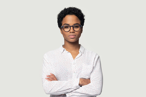 Serious concentrated young african american girl in eyeglasses standing with folded hands, headshot studio portrait. Focused black female professional looking at camera, isolated on grey background.
