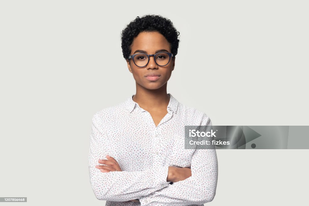 Serious concentrated young african american girl standing with folded hands. Serious concentrated young african american girl in eyeglasses standing with folded hands, headshot studio portrait. Focused black female professional looking at camera, isolated on grey background. Women Stock Photo